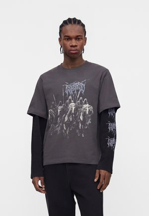 Νεαρός με πλεξούδες φοράει στρωμένα μαύρα και γκρι μακρυμάνικα και κοντομάνικα πουκάμισα με γραφικό σχέδιο καβαλάρηδων με κουκούλες πάνω σε άλογα.