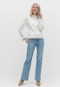Weißer langärmeliger Pullover mit hohem Kragen und taupefarbenen Akzenten, kombiniert mit hellblauen geraden Jeans und beigefarbenen Stiefeletten.