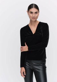 Svart långärmad topp i mjuk sammet med en draperad V-ringning, med en knutdetalj fram, kombinerad med svarta byxor i konstläder.