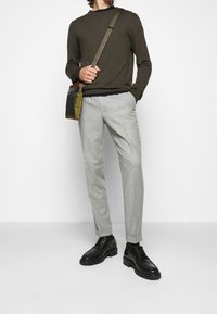 Uomo che indossa un maglione verde oliva, pantaloni grigi chiari su misura, scarpe in pelle nere e una borsa a tracolla camouflage, in piedi davanti a uno sfondo bianco.