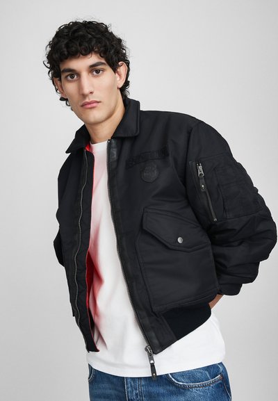 Schwarze Bomberjacke mit Reißverschluss vorne, Seitentaschen und einem Schulterpatch. Mit geripptem Bund und kontrastierendem Innenfutter. Weicher Stoff.