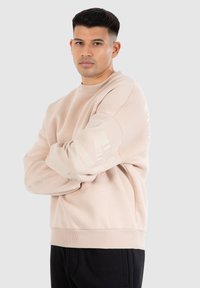 Sweatshirt beige clair avec un col et des poignets côtelés, arborant un motif de logo subtil sur les manches. Tissu doux et texturé.