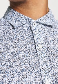 Kortärmad skjorta med vit bakgrund, prydd med blå blommönster, spetsig krage och vita knappar. Mjuk, strukturerad tyg.