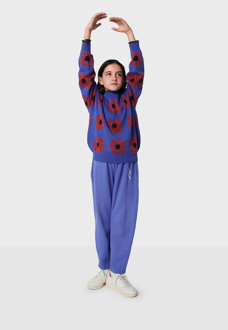 Blå, stickad tröja med röda blommönster, kombinerad med lösa blå sweatpants och vita sneakers. Armarna uppe, stående mot en ljus bakgrund.