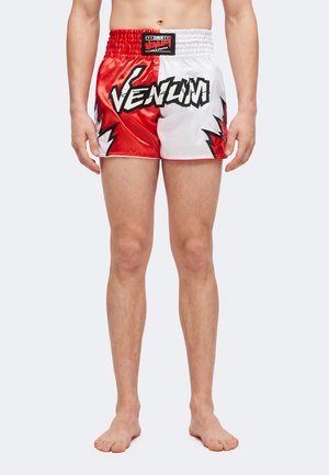 Homme portant un short de Muay Thai rouge et blanc de la marque Venum avec ceinture élastique, debout pieds nus sur un fond uni.