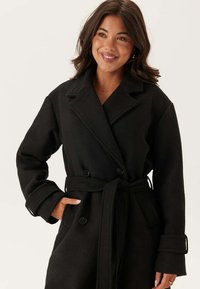 Cappotto in misto lana nero con design doppiopetto, ampi risvolti, vita con cintura e tasche laterali. Texture morbida e vestibilità oversize.