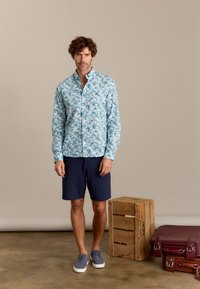 Lichtblauw overhemd met knopen en een bloemenpatroon, gecombineerd met marineblauwe shorts en grijze schoenen. Houten kist en koffers op de achtergrond.