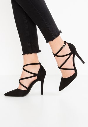 Schwarze High-Heels mit überkreuzten Knöchelriemen kombiniert mit schwarzen Jeans mit fransigem Saum auf einem weißen Hintergrund.