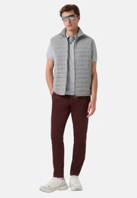 Šedá vypolstrovaná vesta s vysokým límcem, nošená přes světle šedé polokabátové tričko, spárovaná s burgundskými kalhotami a bílými teniskami. Hladká textura, zipové zapínání.