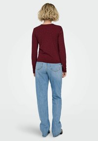 Kobieta z blond kręconymi włosami, ubrana w bordową bluzkę z długim rękawem w drobne kropki oraz jasnoniebieskie dżinsy, stoi tyłem na białym tle.