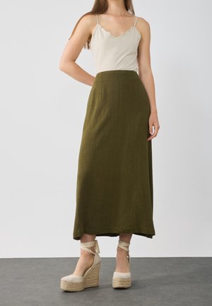 Beige scalloped camisole with thin straps and an olive green, floor-length skirt. Model wearing wedge espadrilles with beige fabric and ankle ties.