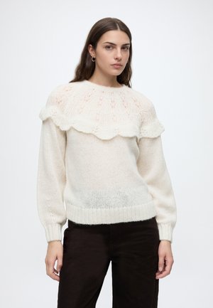 Jeune femme aux cheveux foncés portant un pull en tricot blanc avec un détail en dentelle et un volant sur la poitrine, associé à un pantalon noir.