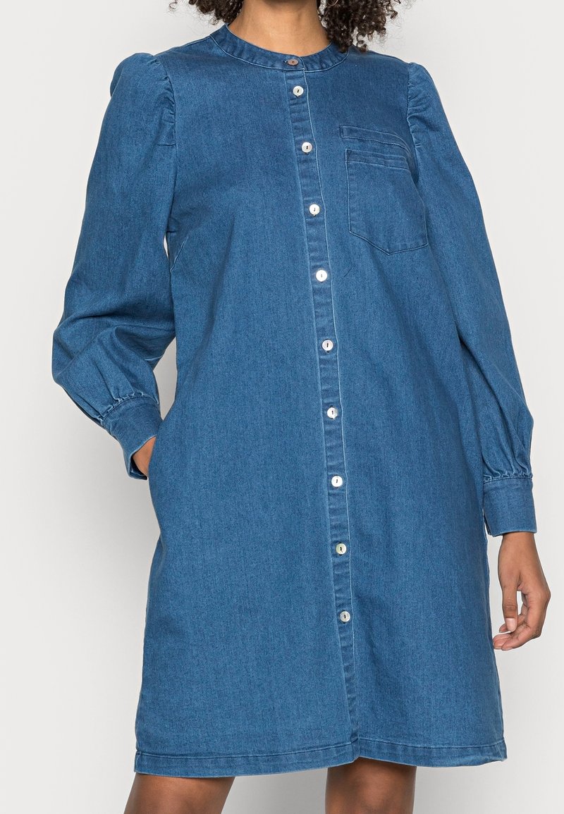 Jeansklänning med rund hals, långa puffärmar, knappstängning fram och en enda bröstficka. Har en medelblå färg och mjuk textur.