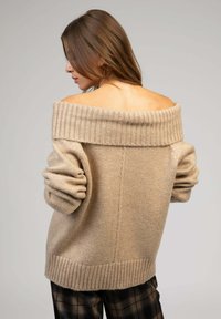 Pull en tricot oversized beige avec un large col côtelé, des épaules tombantes et des poignets et un ourlet côtelés. Texture douce, design décontracté.