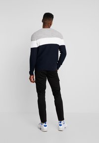 Man wearing a gray, white, and navy color-block sweater with black pants and white sneakers, standing facing away against a plain wall.