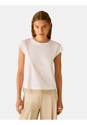 Donna che indossa un top bianco senza maniche con orlo di frange beige e pantaloni beige a gamba larga, accessoriata con gioielli dorati.