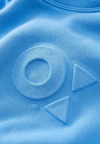 Modrá tkanina s vyvýšenými symboly: kruh a dva trojúhelníky směřující doprava, připomínající tlačítka pro ovládání médií, na měkkém texturovaném povrchu.