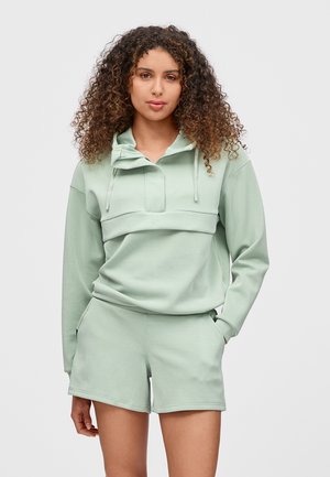 Kvinne med krøllete hår har på seg en blek grønn hettegenser og matchende shorts, står med en hånd i lommen mot en ensfarget lys bakgrunn.