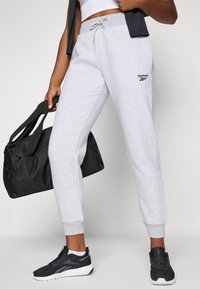 Pantalones de chándal grises de algodón con cintura elástica y cordón, con el logo de Reebok. Combinados con zapatillas deportivas negras y un bolso de deporte negro.