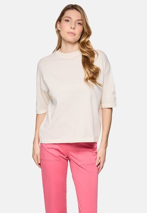 Mujer con cabello largo y ondulado, vestida con una blusa suelta de manga corta color blanco hueso y pantalones de color rosa brillante, de pie contra un fondo claro y liso.