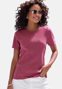 Maroon short-sleeve shirt with a textured diamond pattern, round neckline, and relaxed fit. Worn with white pants and accessorized with earrings.