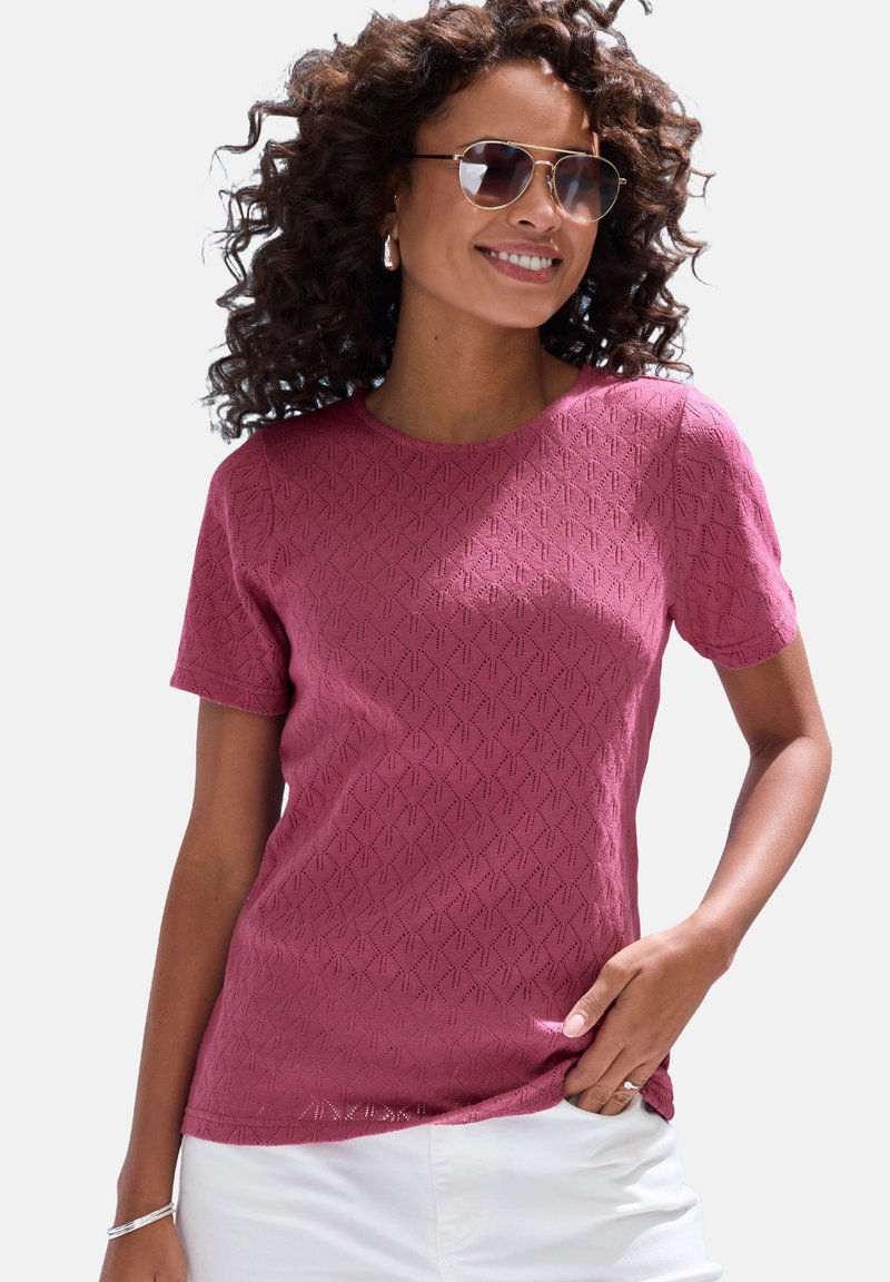 Maroon short-sleeve shirt with a textured diamond pattern, round neckline, and relaxed fit. Worn with white pants and accessorized with earrings.