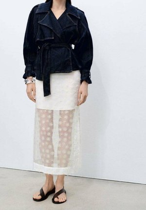 Personne portant une veste noire ceinturée, une jupe blanche transparente à couches avec des motifs floraux, et des sandales noires à bouts ouverts, debout contre un mur uni.