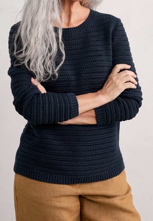 Frau mit langen grauen Haaren verschränkt die Arme, trägt einen dunkelblauen Strickpullover und hellbraune Hosen vor einem neutralen Hintergrund.