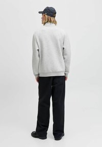 Person i grå sweatshirt, sorte bukser, sorte sko og en mørk kasket, som står med ryggen til foran en ensfarvet lys baggrund.