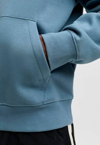 Světle modrá mikina s kapucí, která má přední kapsu, žebrované manžety a spodní lem. Hladká textura s volným střihem, která zdůrazňuje čisté švy.