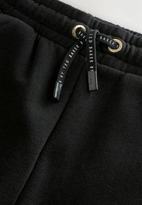 Nærbillede af sort stof fra bukser med elastisk talje, der har sorte snore og prydes af hvid "Ted Baker" tekst samt gyldne metaløjer.