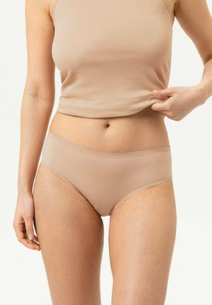 Vrouw draagt naadloze huidkleurige onderkleding en tanktop, houdt de zoom van de top met één hand vast.