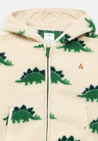 Φούτερ με φερμουάρ από απαλό κρεμ φλις, διακοσμημένο με πράσινα σχέδια δεινοσαύρων, με φερμουάρ σε αντίθετο χρώμα και μικρή κεντητή λεπτομέρεια αρκούδας στο στήθος.