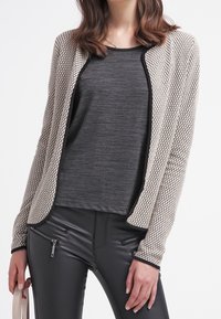 Cárdigan beige texturizado con un patrón de panal, combinado con una blusa negra y pantalones negros ajustados con detalles de cremalleras.