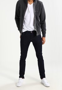 Jean bleu foncé, design ajusté, associé à un sweat à capuche gris zippé et un t-shirt blanc à col en V. Des baskets blanches complètent la tenue.