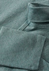 Tessuto color teal con una consistenza morbida. Presenta un colletto alto e una manica con polsino. Le cuciture sono visibili, aggiungendo dettagli al design complessivo.