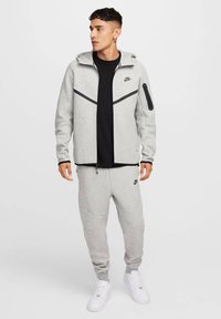 Pilkas Nike susegamas džemperis su juodais akcentais, derinamas su pilkais džinsais. turi didelę kapucę, kišenes ir glotnią tekstūrą.