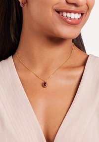 Gouden ketting met een ovale hanger, voorzien van een donkerrode edelsteen omringd door kleinere stenen, op een gestructureerde lichte stof.