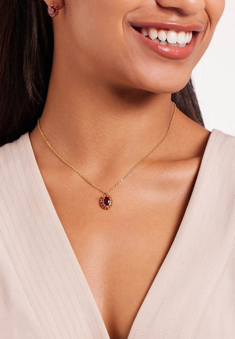 Gouden ketting met een ovale hanger, voorzien van een donkerrode edelsteen omringd door kleinere stenen, op een gestructureerde lichte stof.