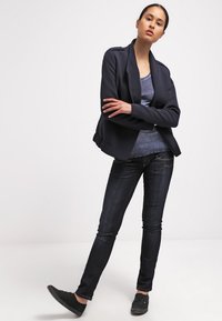 Blazer navy con solapas afiladas, superpuesto a una blusa azul oscura, combinado con jeans de denim oscuro y zapatos negros de tipo slip-on.