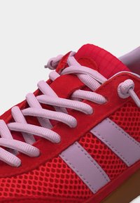 Chaussure de sport rouge avec des lacets roses, une tige en mesh, deux rayures rose clair sur le côté et une semelle en gomme.