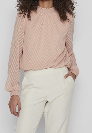 Vrouw draagt een blushroze textuurblouse met lange mouwen en chevronpatroon, gecombineerd met een high-waisted crèmekleurige broek, één hand in de zak.