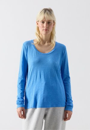 Mlada oseba z blond lasmi, oblečena v svetlo modro majico z dolgimi rokavi in svetlo sive hlače, stoji pred enobarvnim belim ozadjem.