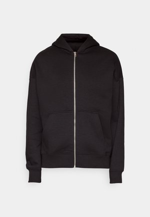 Personne portant un pantalon beige, des baskets noires et blanches, et un sweat à capuche noir par-dessus un t-shirt marron, debout de profil sur un fond uni.