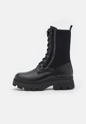 Bota de combate de cuero negro y tela con suela gruesa, cremallera lateral y cordones delanteros, con la marca Calvin Klein Jeans en la tira de la cremallera.