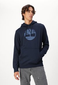 Marineblauwe katoenen hoodie met een cirkelvormig boomlogo in donkerblauw, voorzien van een buidelzak en een trekkoordcapuchon, met textuurstof en lange mouwen.
