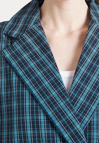 Modrý a černý kostičkovaný blazer s širokým límcem, který má čtvercový vzor a hladkou texturu. Nosit přes bílý top.