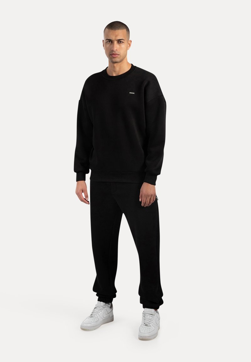 Černá mikina s dlouhými rukávy, žebrovanými manžetami a kulatým výstřihem. V kombinaci s černými tepláky a bílými teniskami. Jednoduchý, minimalistický design.