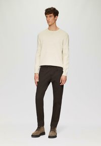 Beige Strickpullover mit Rundhalsausschnitt, kombiniert mit dunkelbraunen, schmal geschnittenen Hosen. Das Model trägt hellbraune Schnürstiefel. Einfaches, strukturiertes Design.