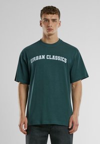 Zöld pamut póló laza szabással, kerek nyakkal és rövid ujjal. A mellkason vastag fehér betűkkel van feltüntetve az "URBAN CLASSICS" felirat.